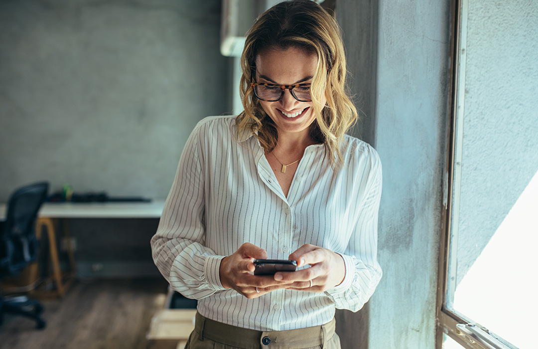 photo-business_woman_mobile_banking-1080x700 A business woman smiles while looking at her mobile phone